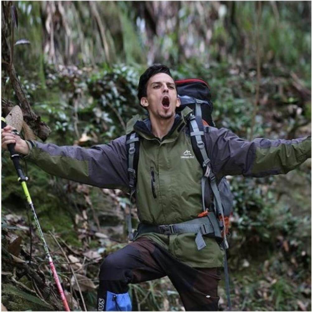 A man outdoors trekking with his arms wide and mouth open as if he's shouting. He is carrying a carbon fiber hiking pole in his right hand and carrying a backpack.