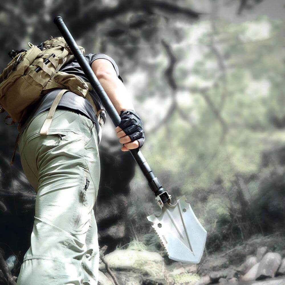 A man outdoors wearing a backpack trekking while holding the multi-tool shovel.